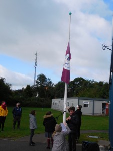 Múinteoir Sandra agus páistí i Rang a 3 ag ardú brat nua na scoile, le Tadhg ó Rang a 6 ag seinm Amhrán na bhFiann ar an bhfeadóg stáin. 